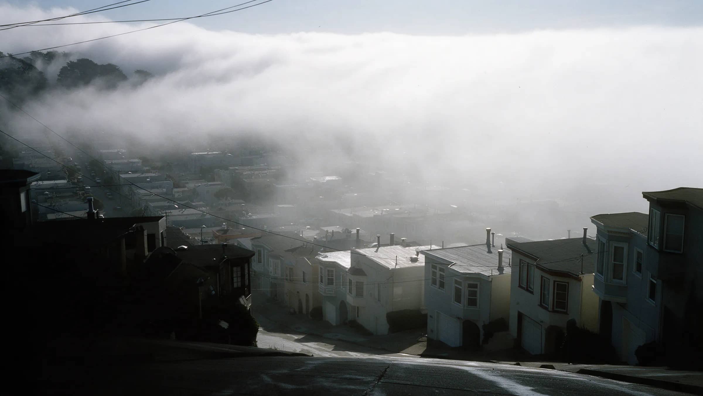 A street of Sunset rowhouses in afternoon fog, garages under the living floors, parked cars beaded with damp and nobody in the frame, San Francisco.