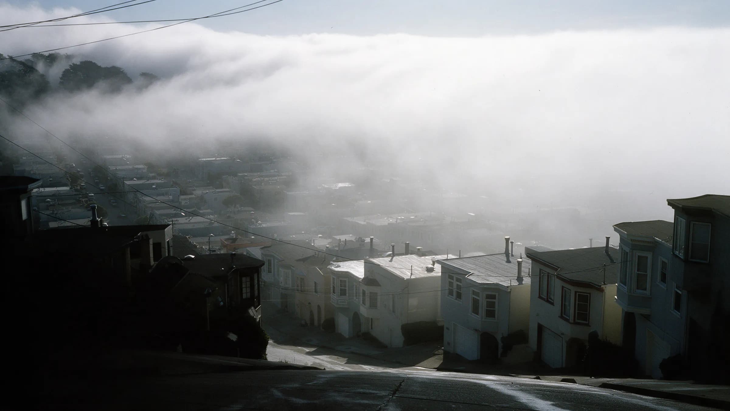 A street of Sunset rowhouses in afternoon fog, garages under the living floors, parked cars beaded with damp and nobody in the frame, San Francisco.